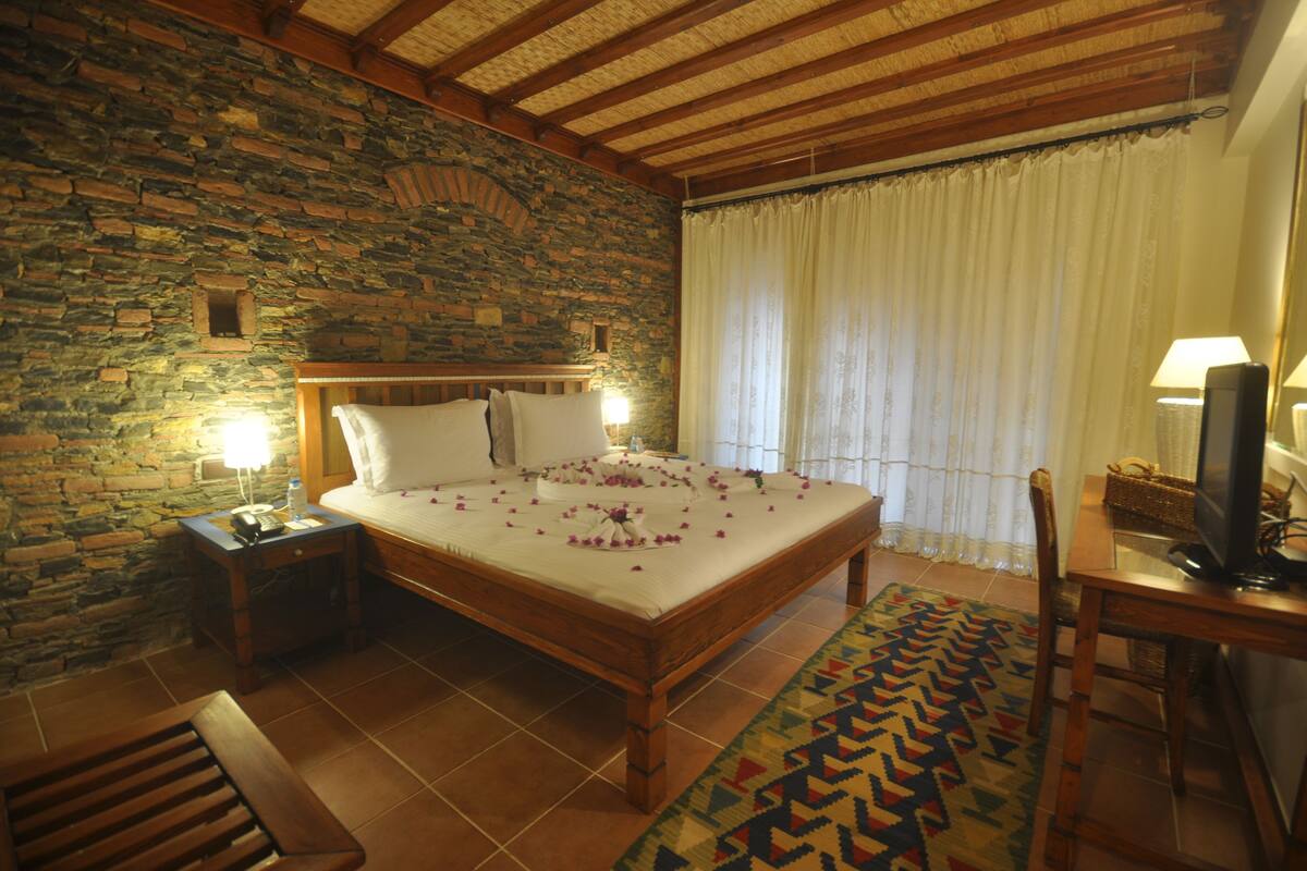 A room with a stone wall, a wooden bed with white linens, a bedside table with a lamp, a television on a wooden stand, and a patterned rug.