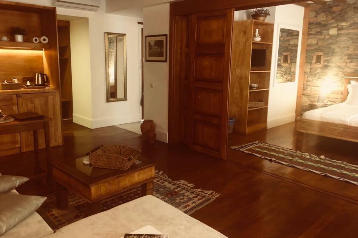 A cozy living room with a wooden coffee table, a sofa, and a patterned rug. There is a wooden door leading to another room and a stone wall in the background.