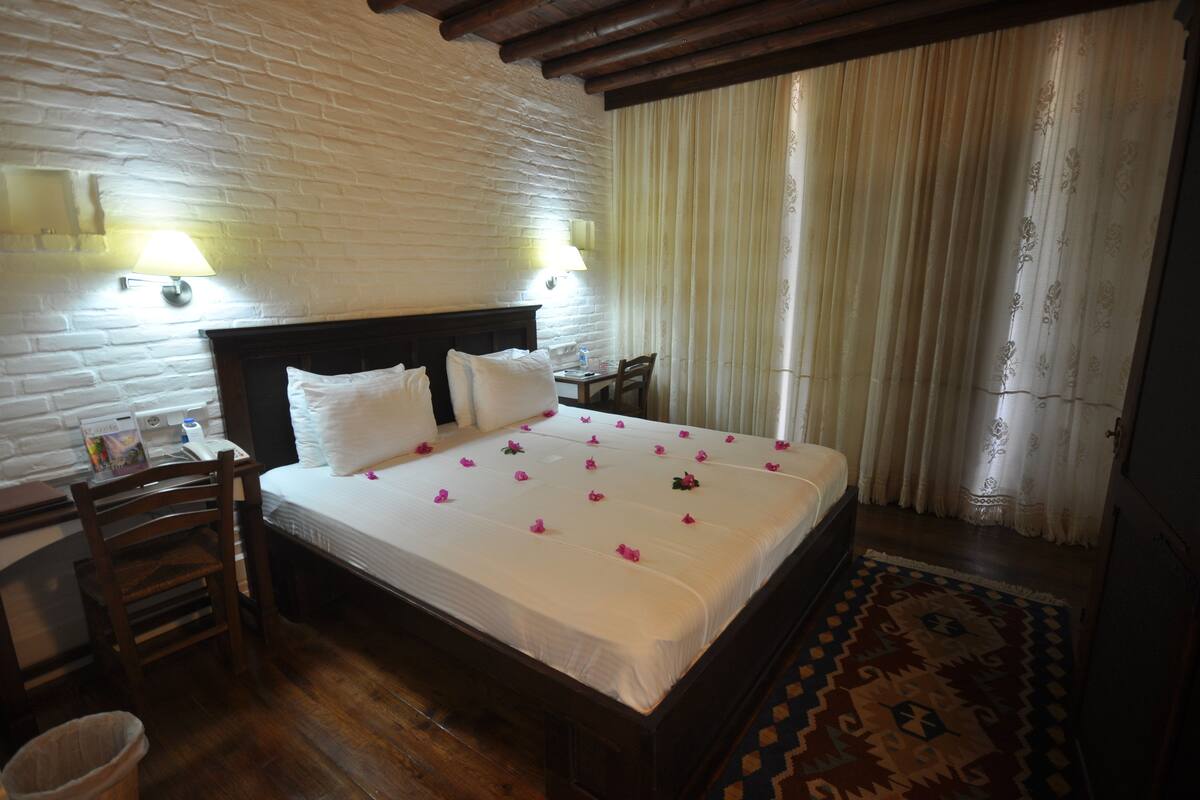 A neatly made bed with white linens and pink flower petals, a wooden nightstand with a lamp, a chair, and a patterned rug.