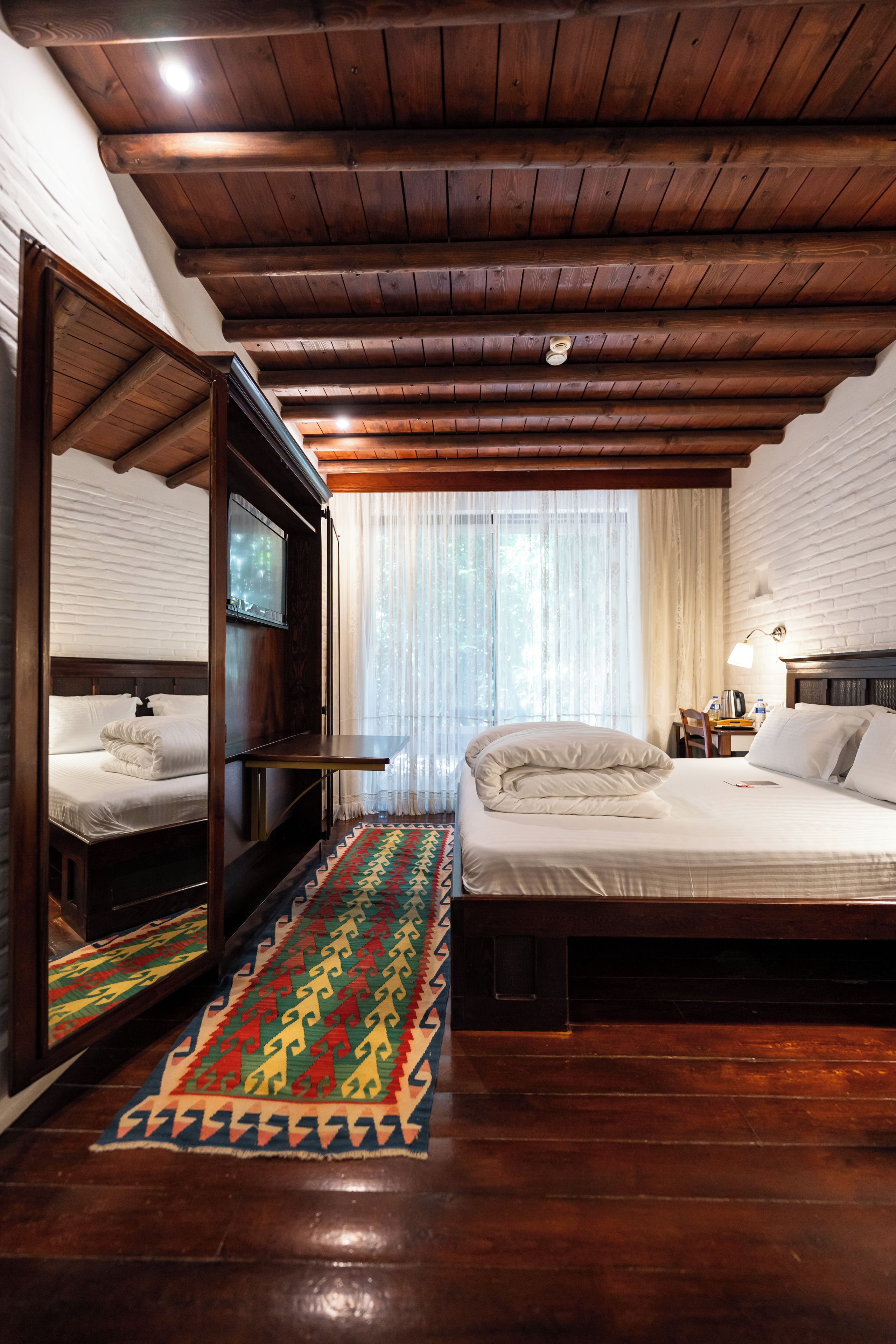 A hotel room with a wooden ceiling, a bed with white bedding, a large mirror, and a colorful rug.