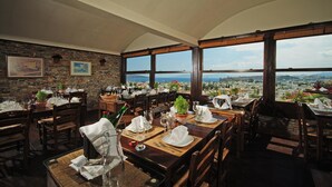 Un restaurante con varias mesas dispuestas para comer, amplios ventanales que ofrecen vistas a la ciudad y una pared de piedra con cuadros enmarcados.