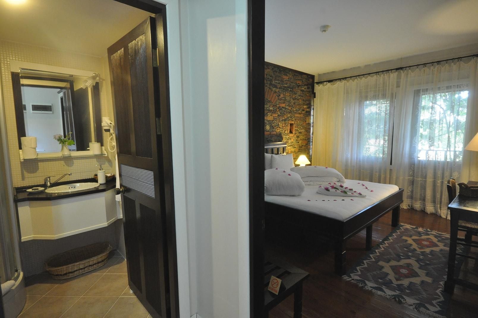A hotel room with a bathroom and a bedroom. The bathroom has a sink, mirror, and towel rack. The bedroom has a bed, nightstand, and a window with curtains.