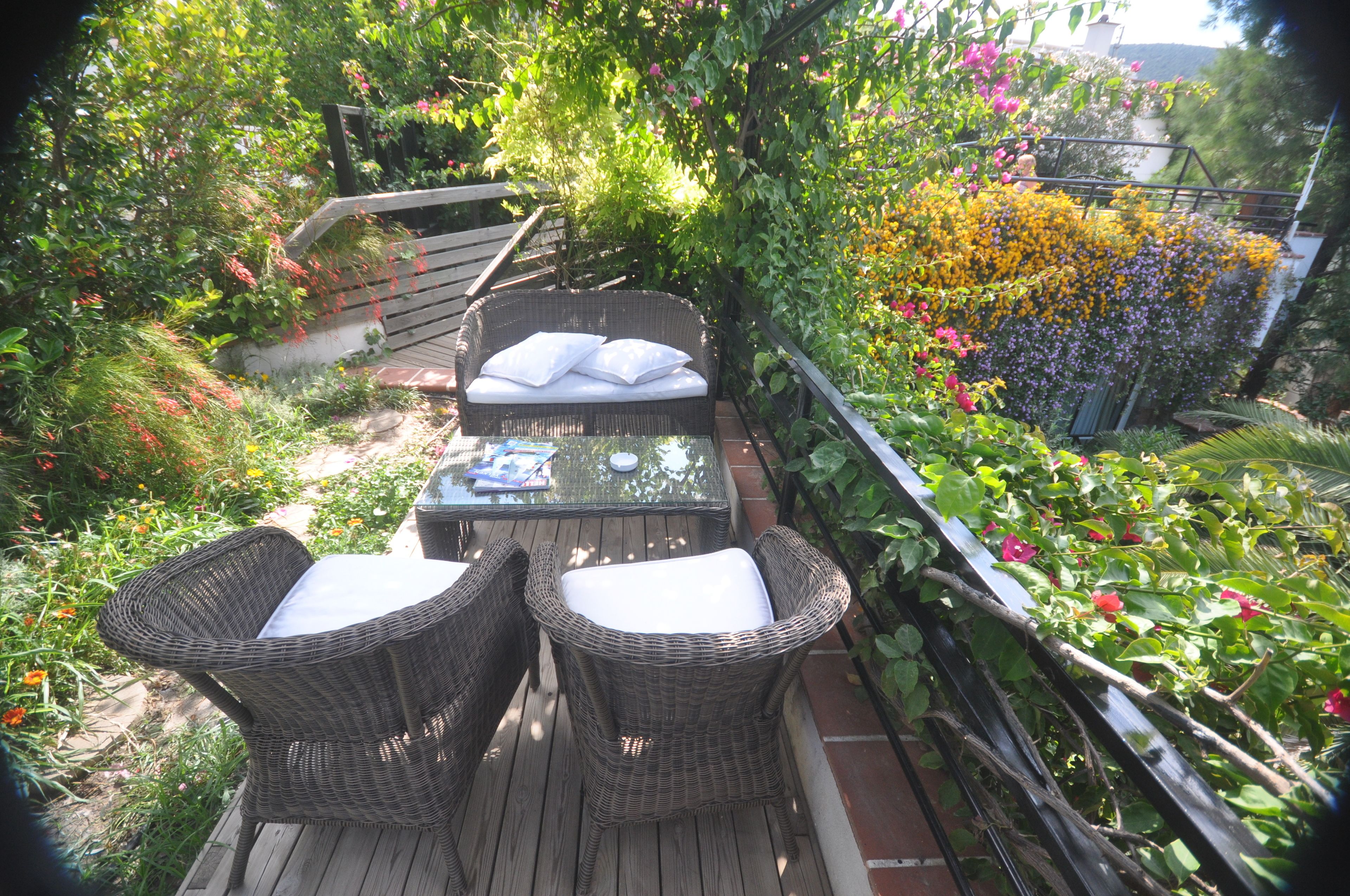 A wooden deck with wicker furniture and a glass table, surrounded by lush greenery and flowering plants.
