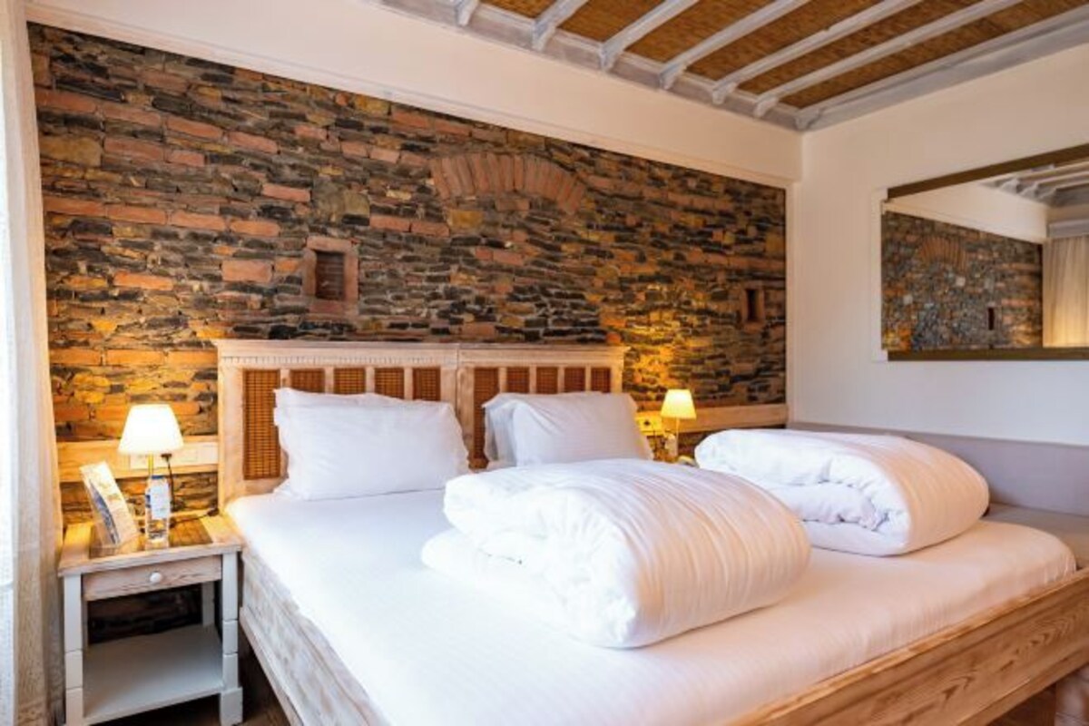 A double bed with white linens, a stone wall, and a wooden ceiling.