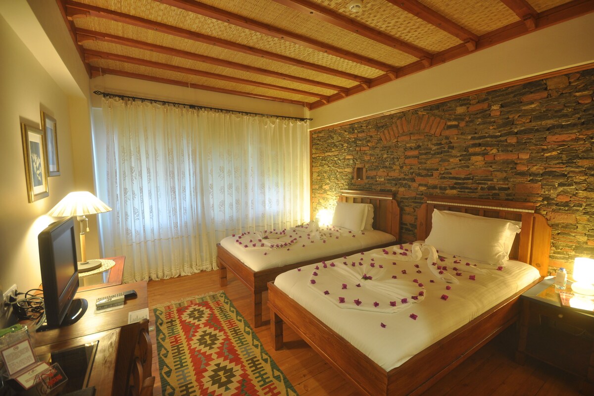 A hotel room with two wooden beds, a stone wall, a wooden ceiling, and a desk with a lamp.