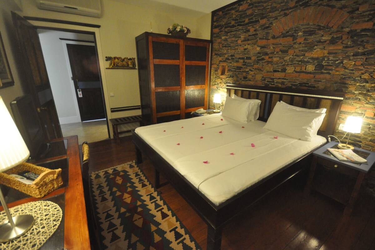 A neatly made bed with white linens and rose petals, a wooden nightstand with a lamp, a wicker chair, and a stone wall.