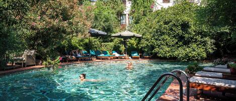 Une piscine à l’eau claire, entourée d’une végétation luxuriante et bordée d’un bâtiment doté de balcons.