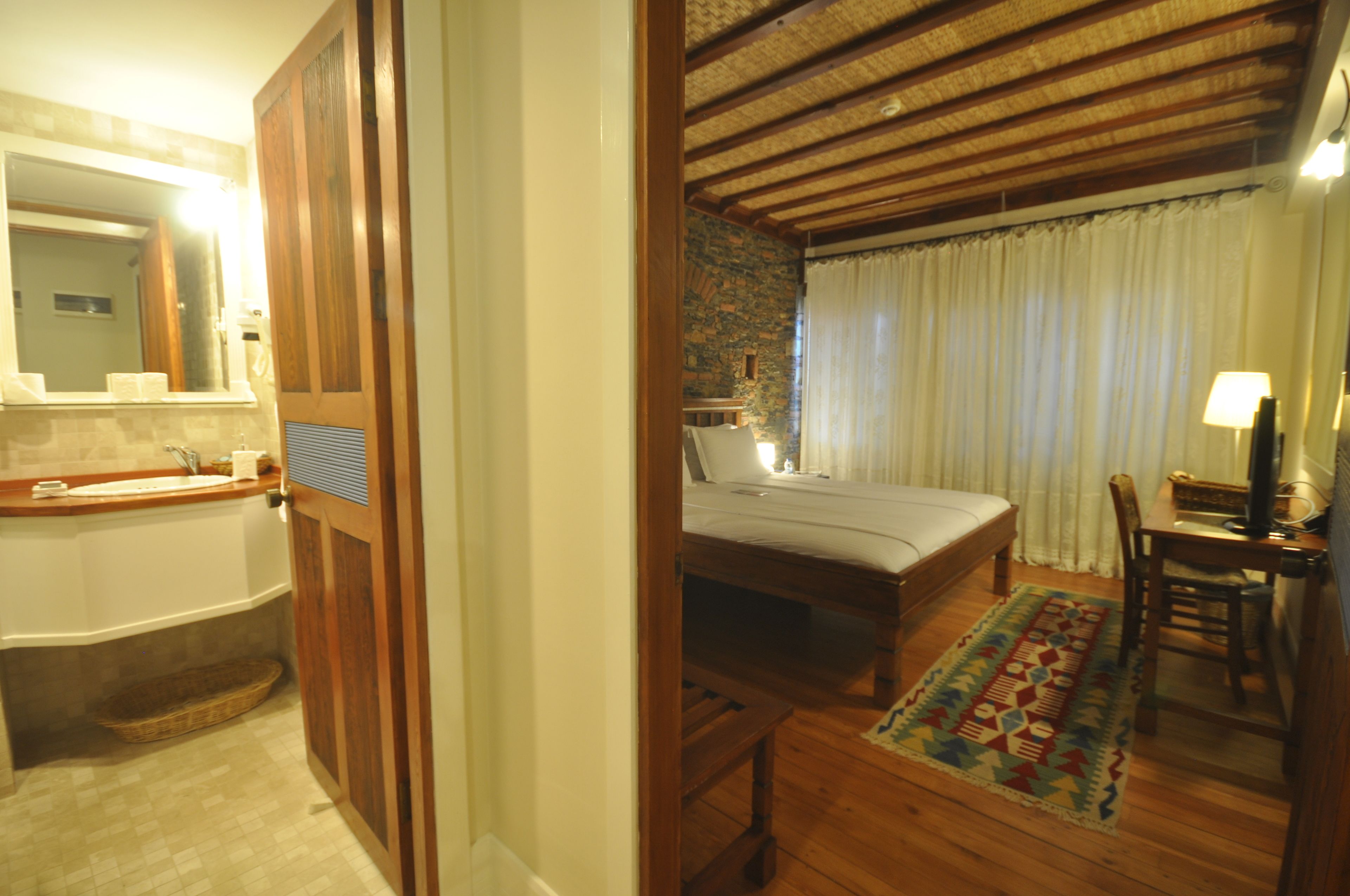 A bedroom with a wooden bed, a desk with a chair, a window with curtains, and a bathroom with a bathtub and sink.
