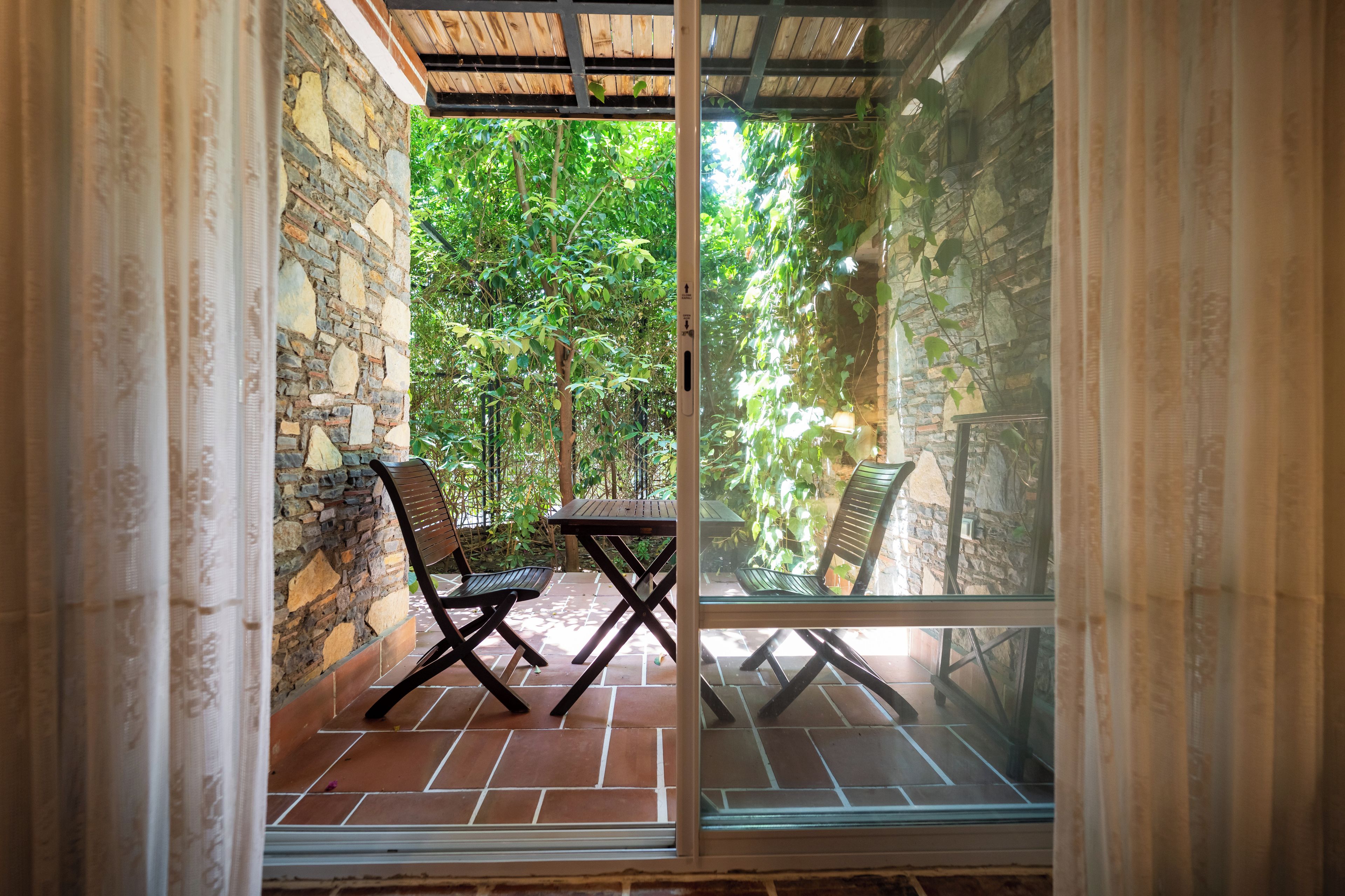 Un patio con una mesa y dos sillas, un muro de piedra y vistas a un jardín exuberante a través de un ventanal.