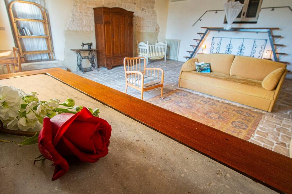 Una stanza con una panca di legno, una rosa rossa, un armadietto di legno, una sedia bianca, un divano beige e un tappeto decorato.