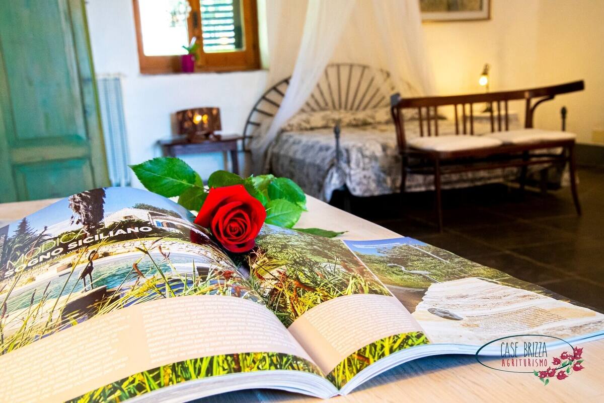 Una stanza con un letto, una panca e un libro con una rosa rossa sopra.