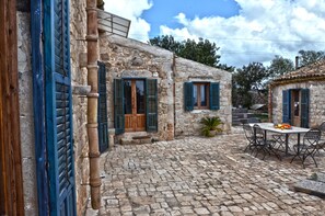 Un edificio de piedra con contraventanas de madera, un patio empedrado y una mesa con sillas.