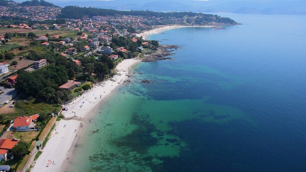 An aerial view of a coastal town with a long sandy beach, clear blue waters, and a backdrop of mountains.