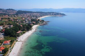Vista aérea de un pueblo costero con una playa extensa de arena, aguas azules cristalinas y montañas al fondo.