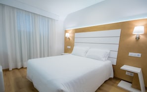 Una habitación de hotel con una cama grande, cortinas blancas, un cabecero de madera y lámparas de noche.