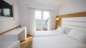 A hotel room with a bed, a desk, a TV, a balcony with a view of the sea, and a window with curtains.