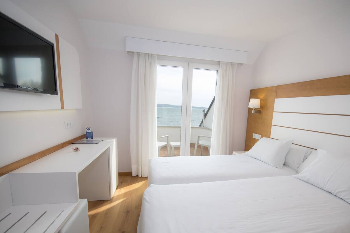 Une chambre d’hôtel avec un lit, un bureau, une télévision, un balcon donnant sur la mer et une fenêtre avec des rideaux.