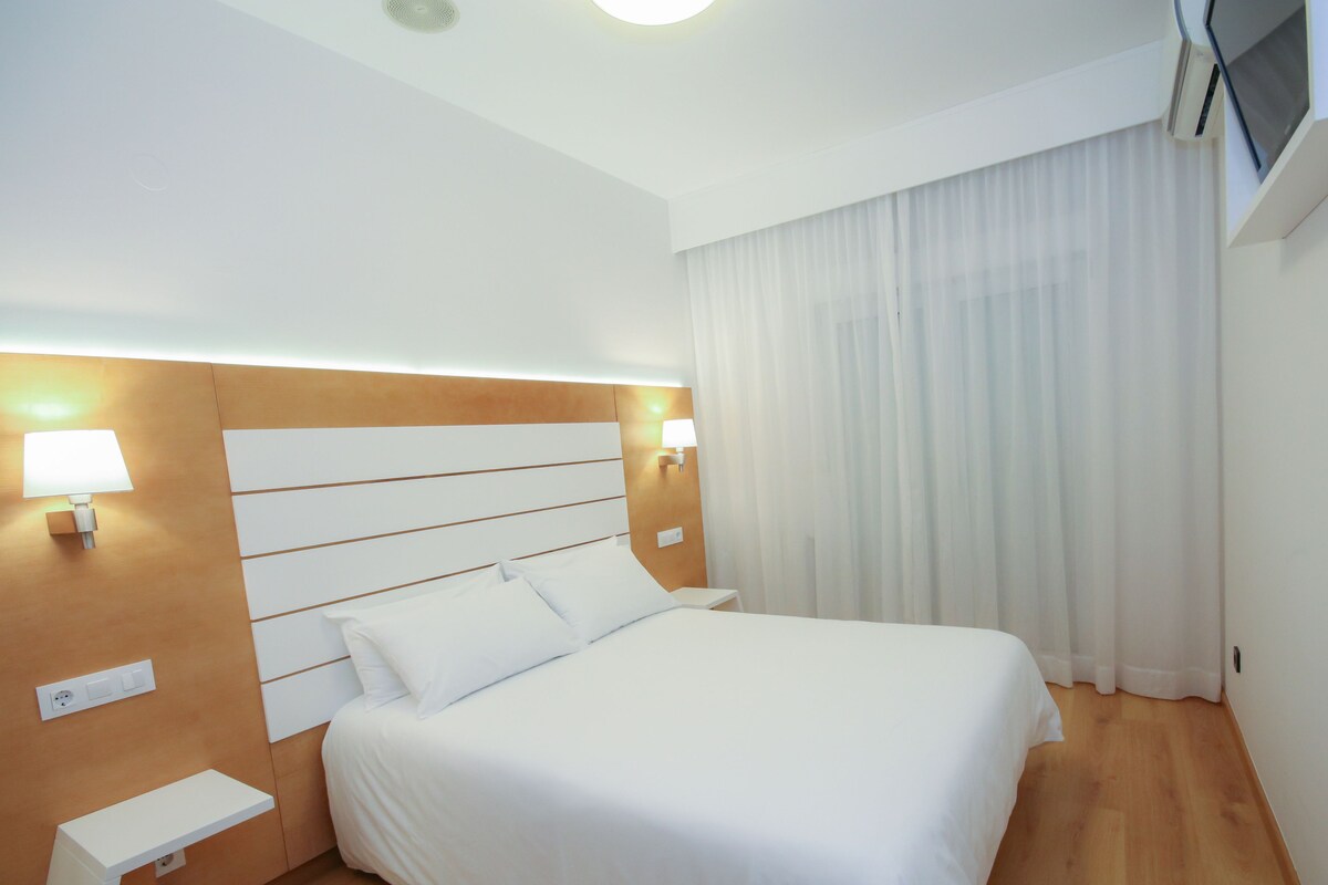 Een hotelkamer met een groot bed, twee bedlampjes, een houten hoofdeinde, een wit nachtkastje en een raam met witte gordijnen.