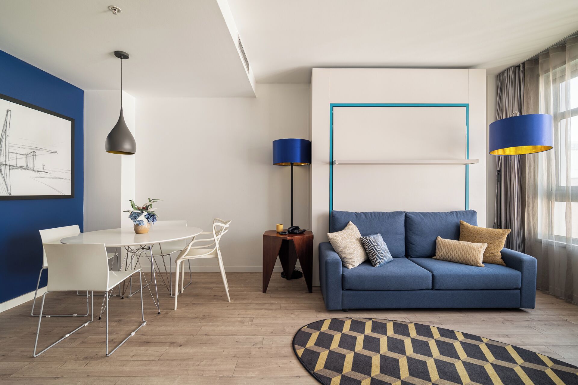 A modern living room with a blue sofa, white dining table, and chairs. There is a floor lamp, a painting on the wall, and a patterned rug.