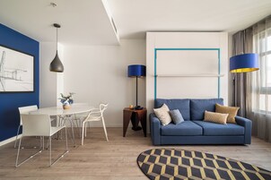 Una sala de estar moderna con un sofá azul, una mesa de comedor blanca y sillas. Hay una lámpara de pie, un cuadro en la pared y una alfombra estampada.