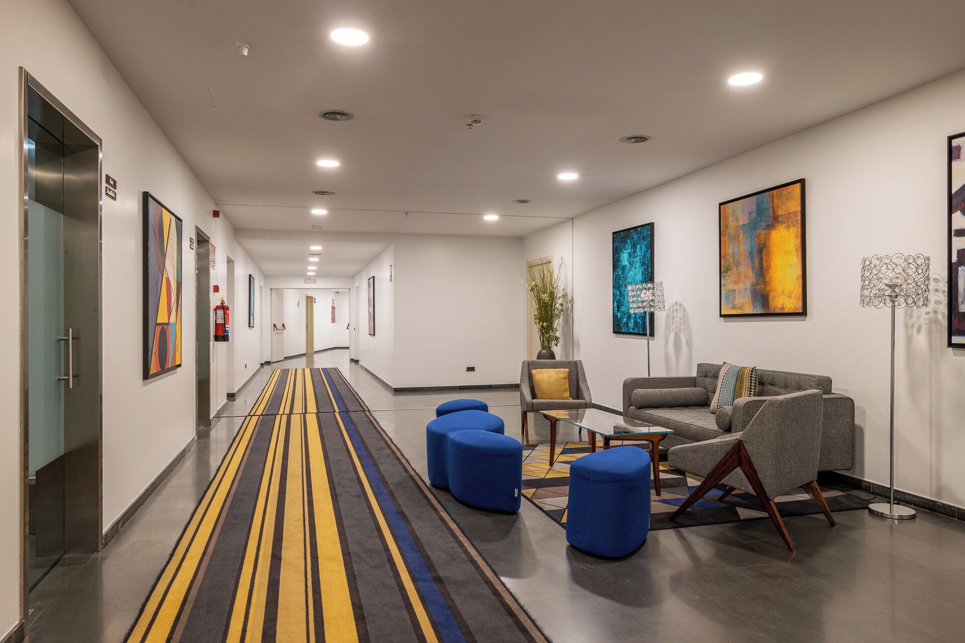 Un moderno vestíbulo de hotel con un largo pasillo, zona de estar con sofás y otomanas, y obras de arte en las paredes.