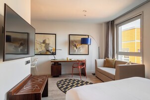 A modern hotel room with a bed, a desk, a chair, and two paintings on the wall.