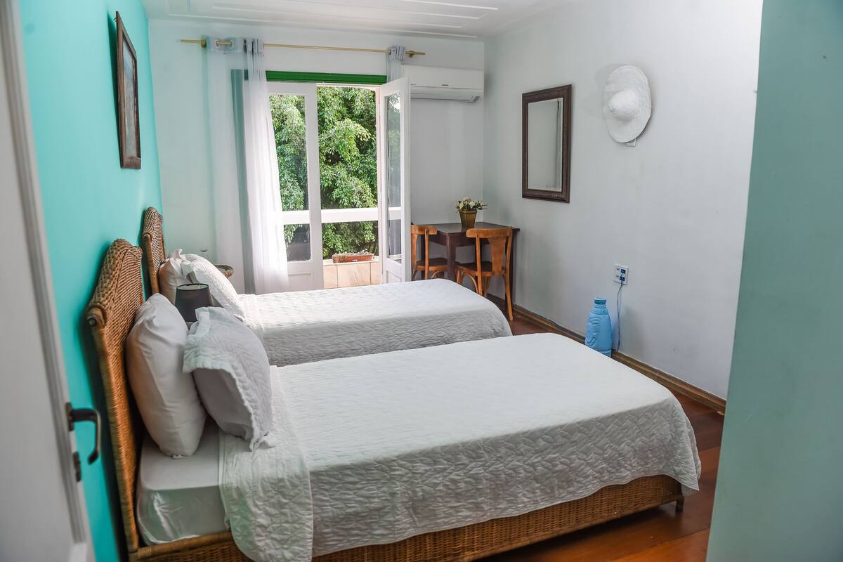 Une chambre avec deux lits, une petite table sur laquelle est posée une plante, un miroir et une fenêtre donnant sur un paysage verdoyant.