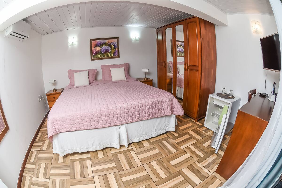 Une chambre avec un grand lit, un plancher en bois, une armoire, une table de chevet, un téléviseur et un luminaire fixé au mur.