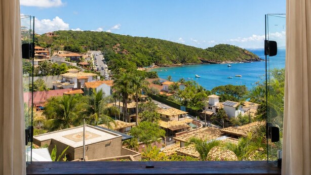 A room with a large arched window overlooking a coastal town, a view of the sea, and a clear sky. - Casagrande Hotel Boutique (Búzios)