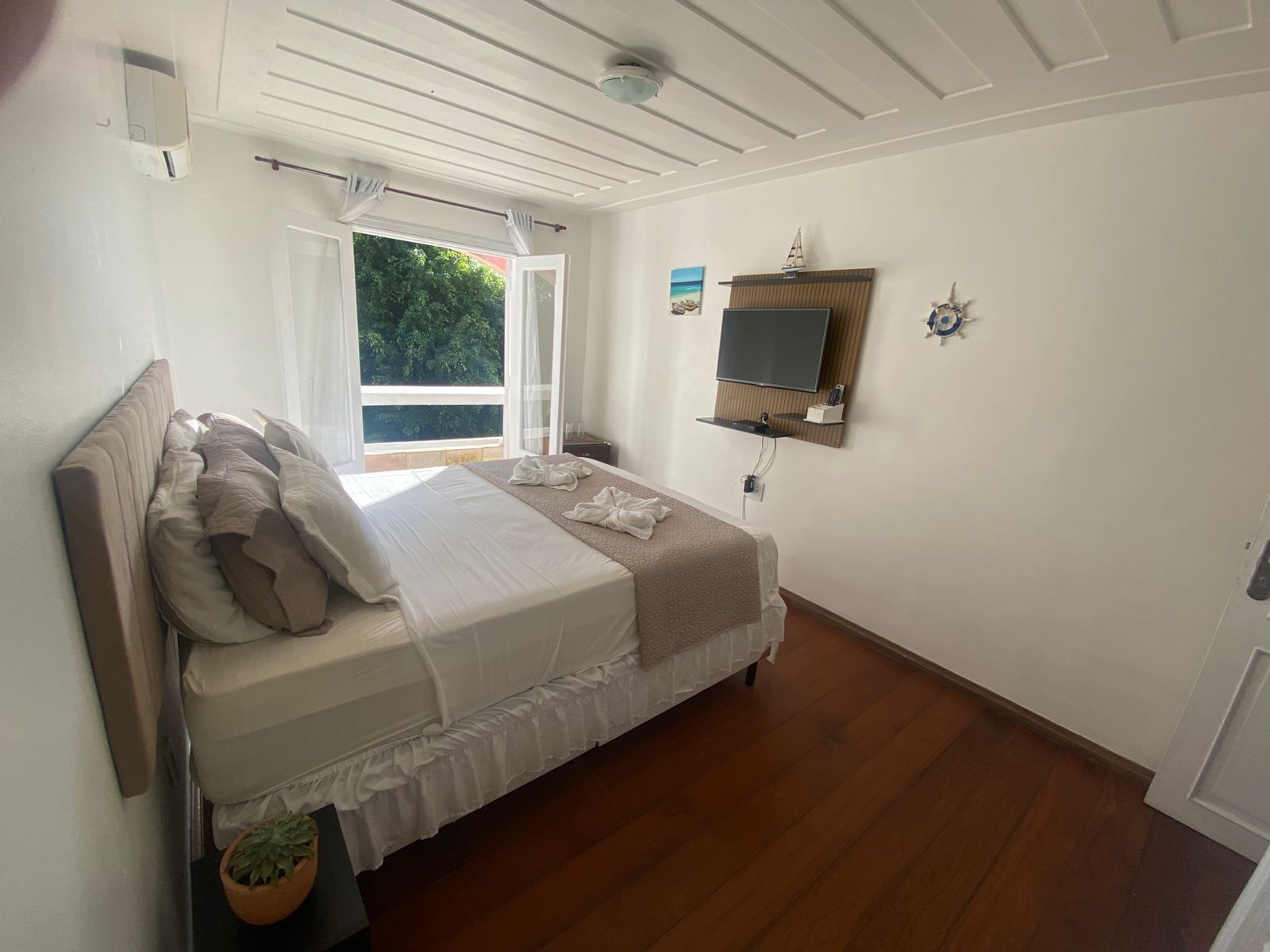 Un dormitori amb un llit, un televisor fixat a la paret, una finestra amb cortines i una petita estant.