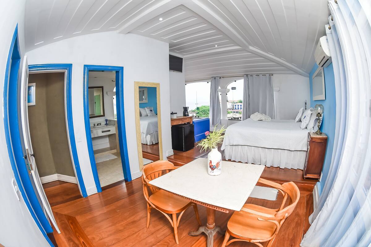 Une chambre d’hôtel comprenant un lit, une table à manger avec des chaises, un miroir, une petite table avec un vase et une fenêtre avec des rideaux.