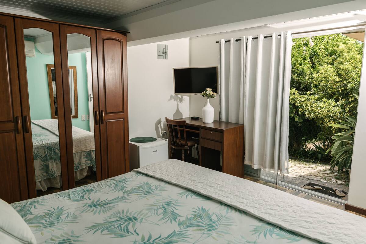 Une chambre à coucher comprenant un lit, un bureau, une chaise, un téléviseur et donnant sur un espace verdoyant.