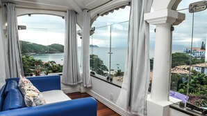 A room with a blue sofa, large windows with curtains, and a view of a coastal area. - Casagrande Hotel Boutique (Búzios)