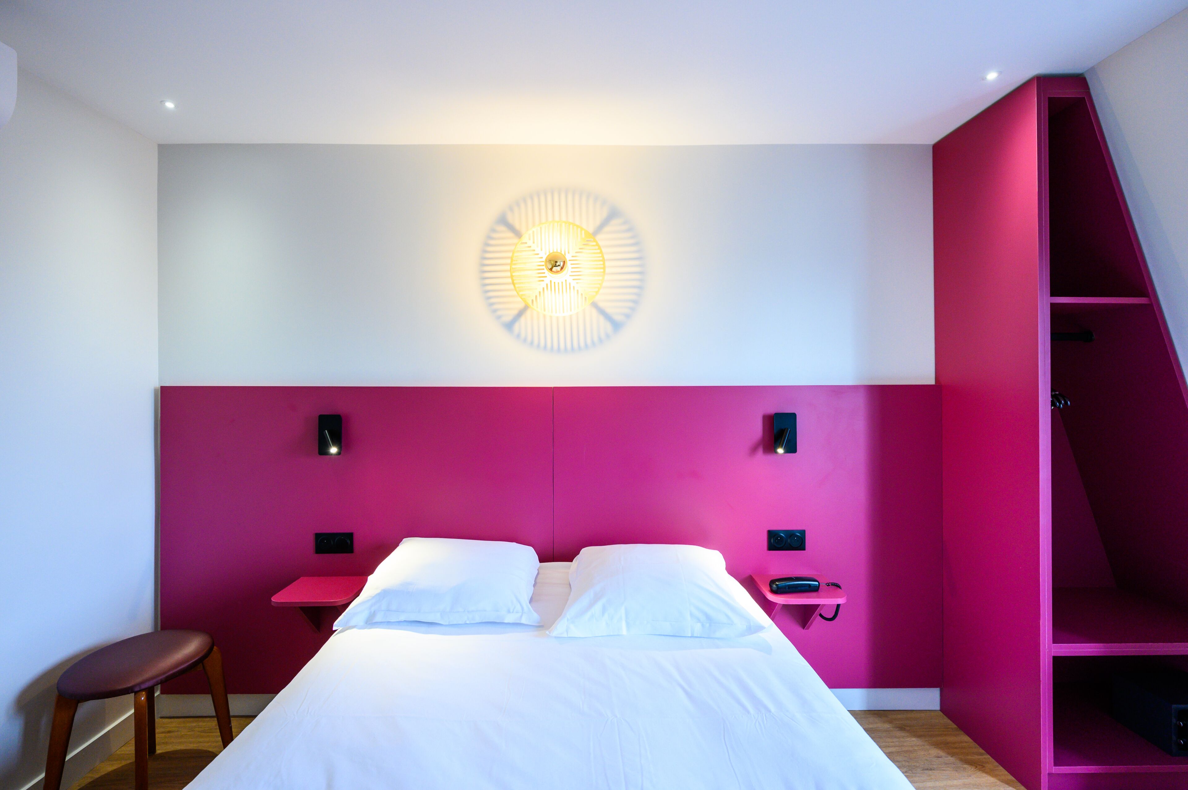 A modern bedroom with a pink accent wall, a bed with white linens, a small bedside table with a lamp, and a wall-mounted light fixture.