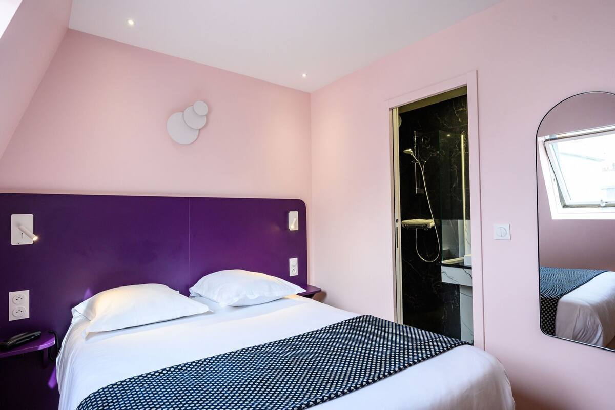 A hotel room with a bed, a shower, a mirror, and a window.