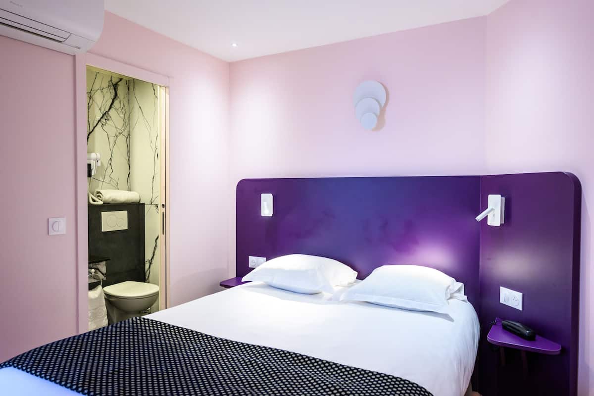 A hotel room with a bed, a nightstand, a bathroom with a toilet, and a wall-mounted light fixture.