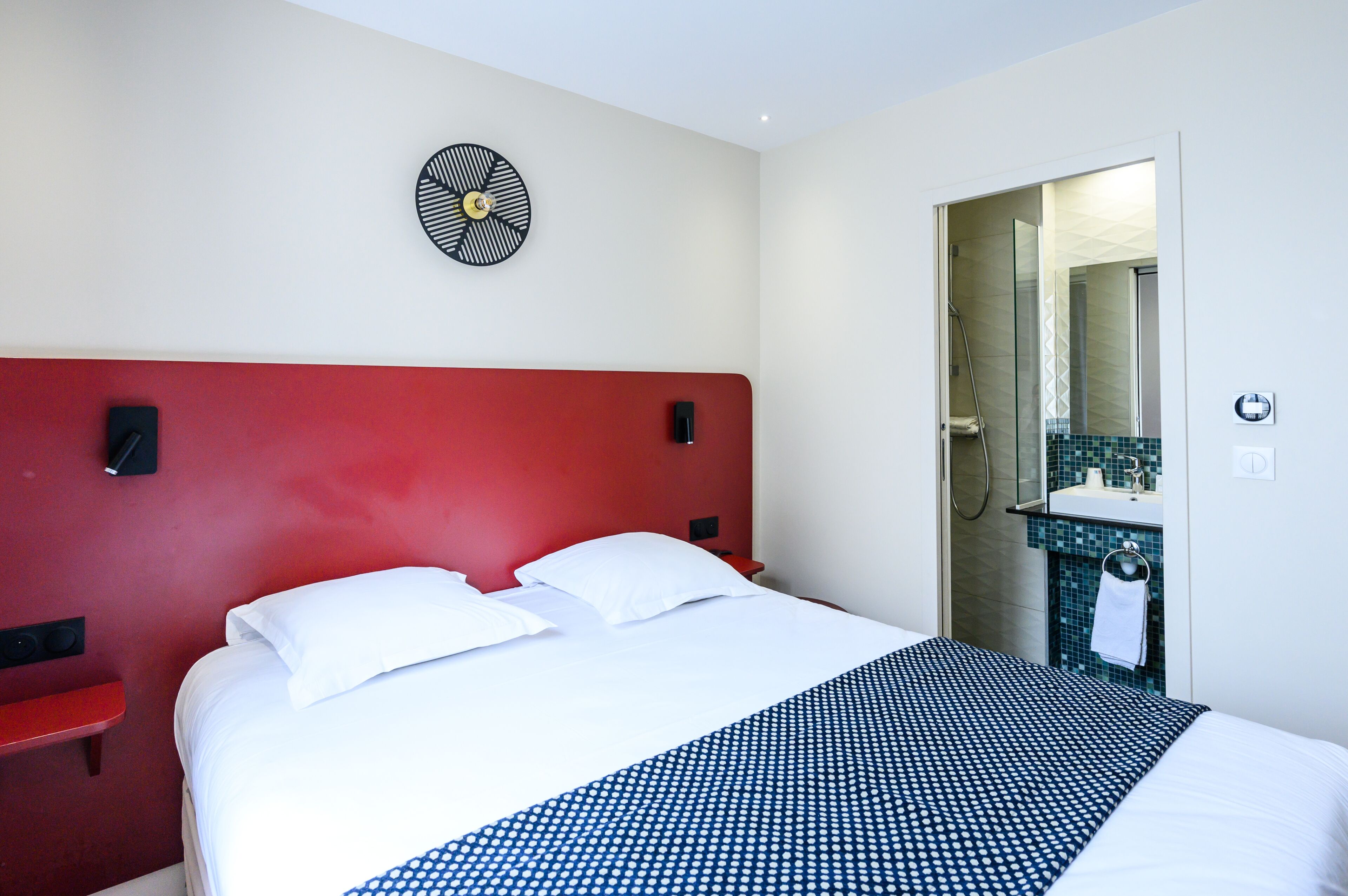 A hotel room with a red headboard, a bed with white linens, a blue and white patterned bedspread, a wall clock, and a bathroom visible through an open door.