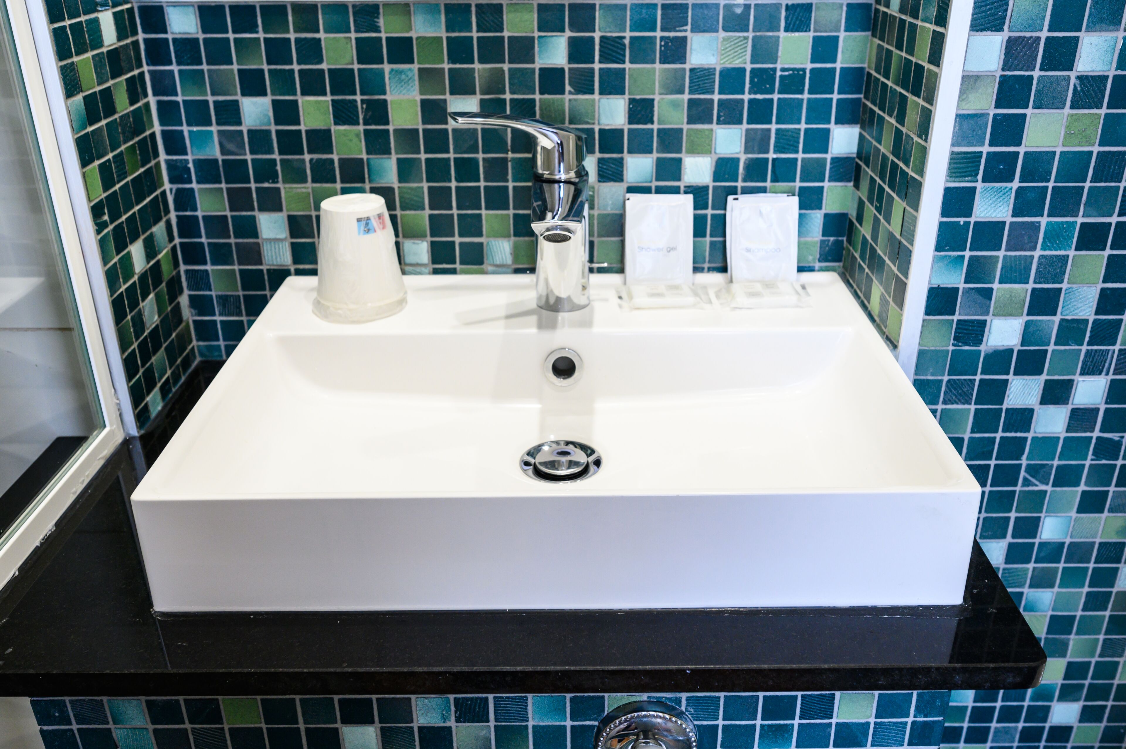A white rectangular bathroom sink with a chrome faucet, a soap dispenser, and a hand soap container. The sink is set against a tiled wall with blue and green mosaic tiles.