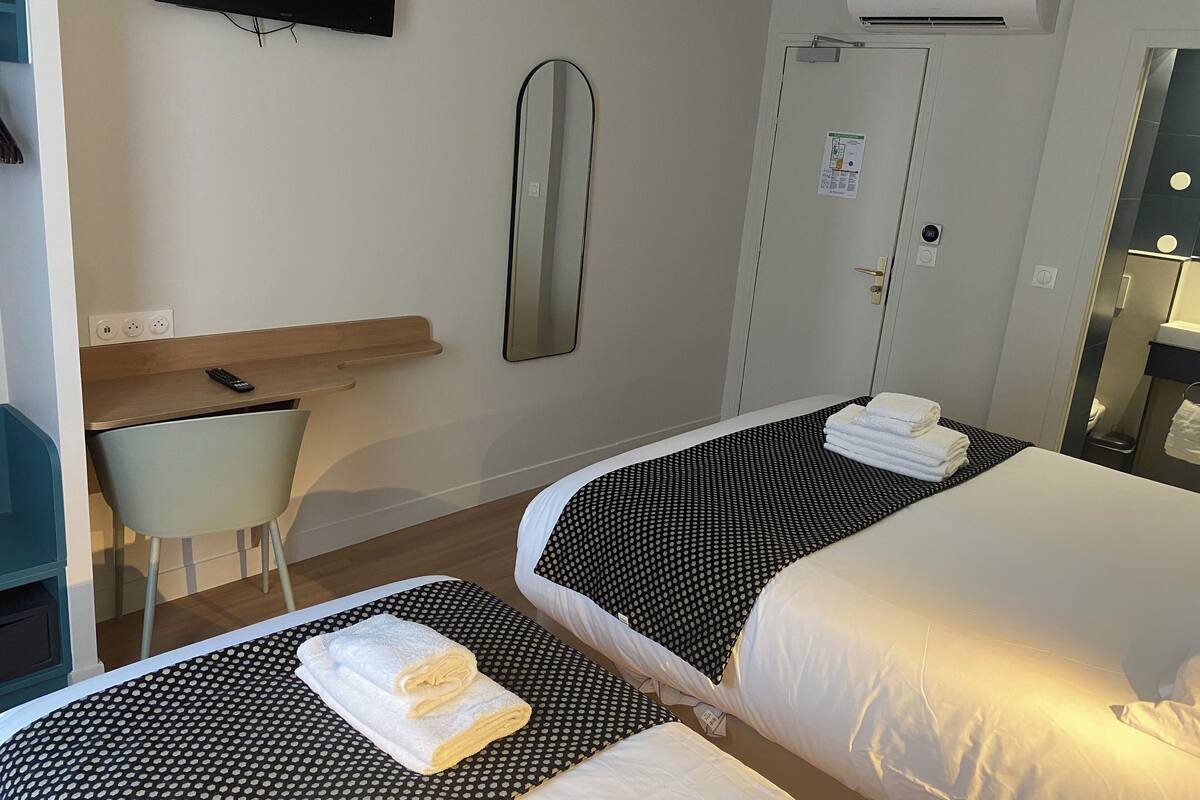 A hotel room with two beds, a desk, a chair, a TV, and an air conditioning unit.