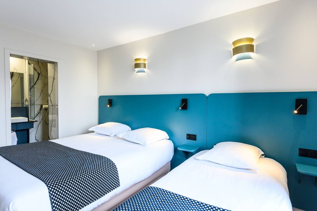 A hotel room with two beds, a bathroom with a mirror, and wall-mounted lighting.