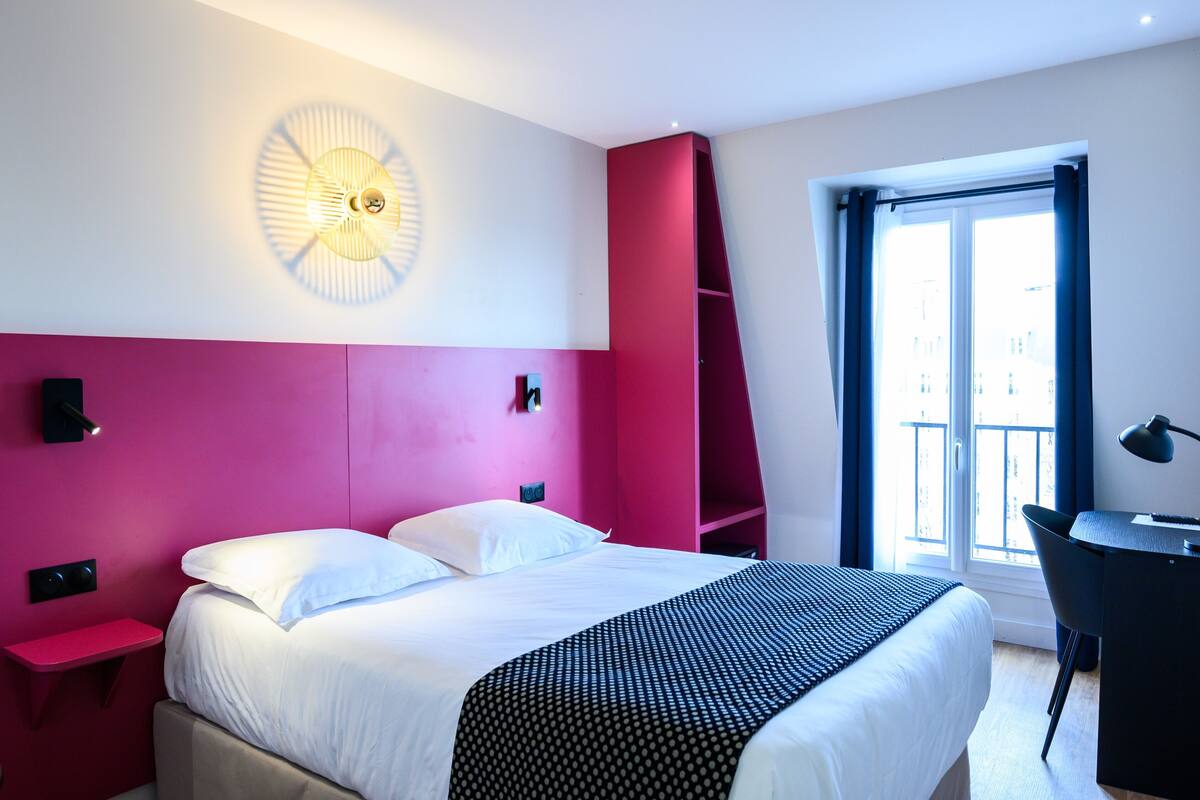 A modern hotel room with a bed, a desk, a chair, a standing shelf, and a window with a view.