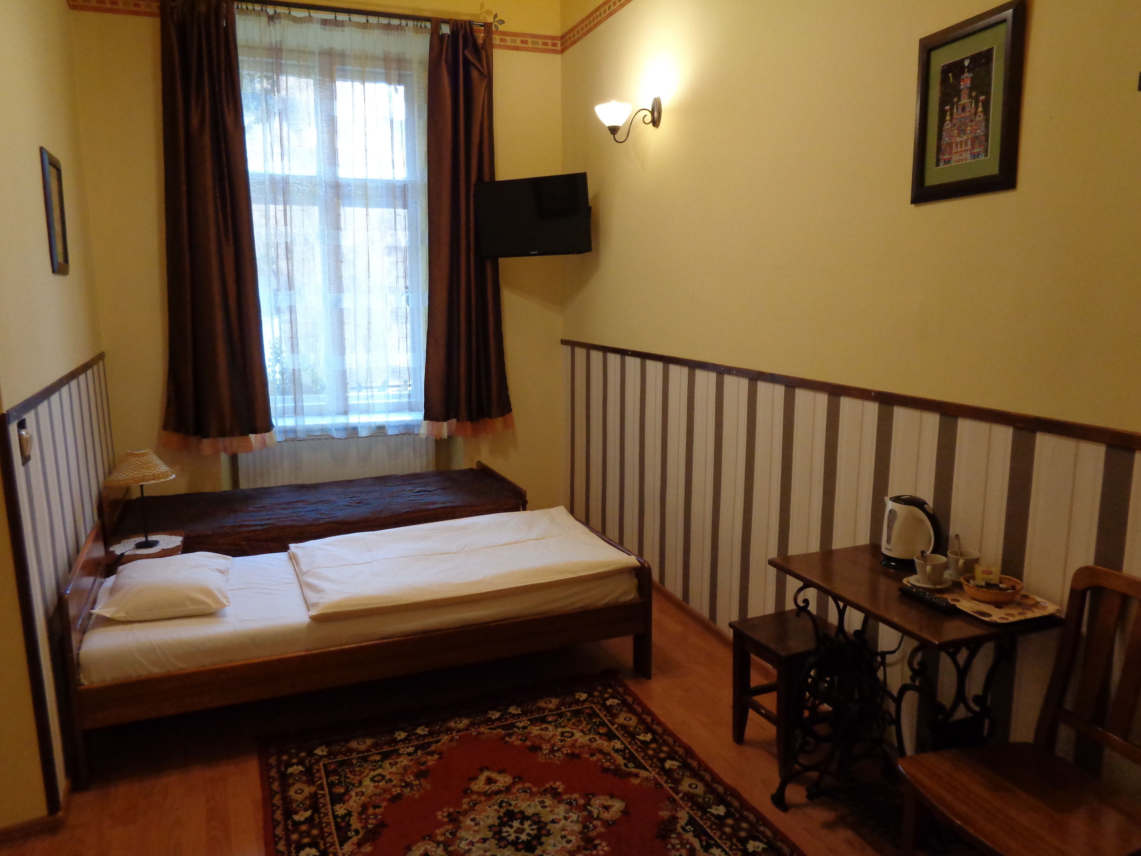 A room with a bed, a small table with a kettle, a chair, a television mounted on the wall, and a window with curtains.