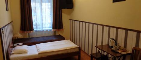 A room with a bed, a small table with a kettle, a chair, a television mounted on the wall, and a window with curtains.