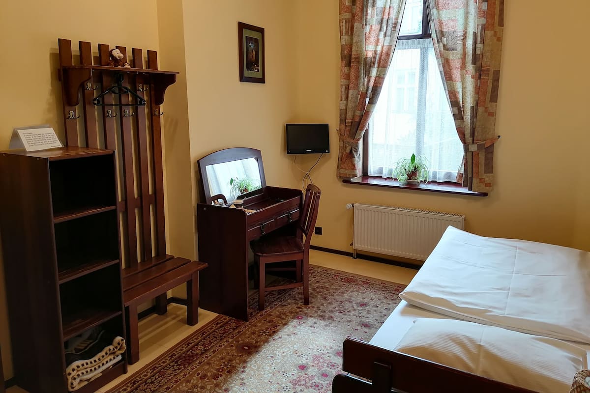 A room with a bed, a desk with a computer, a chair, a radiator, a window with curtains, and a shelf with a sign.