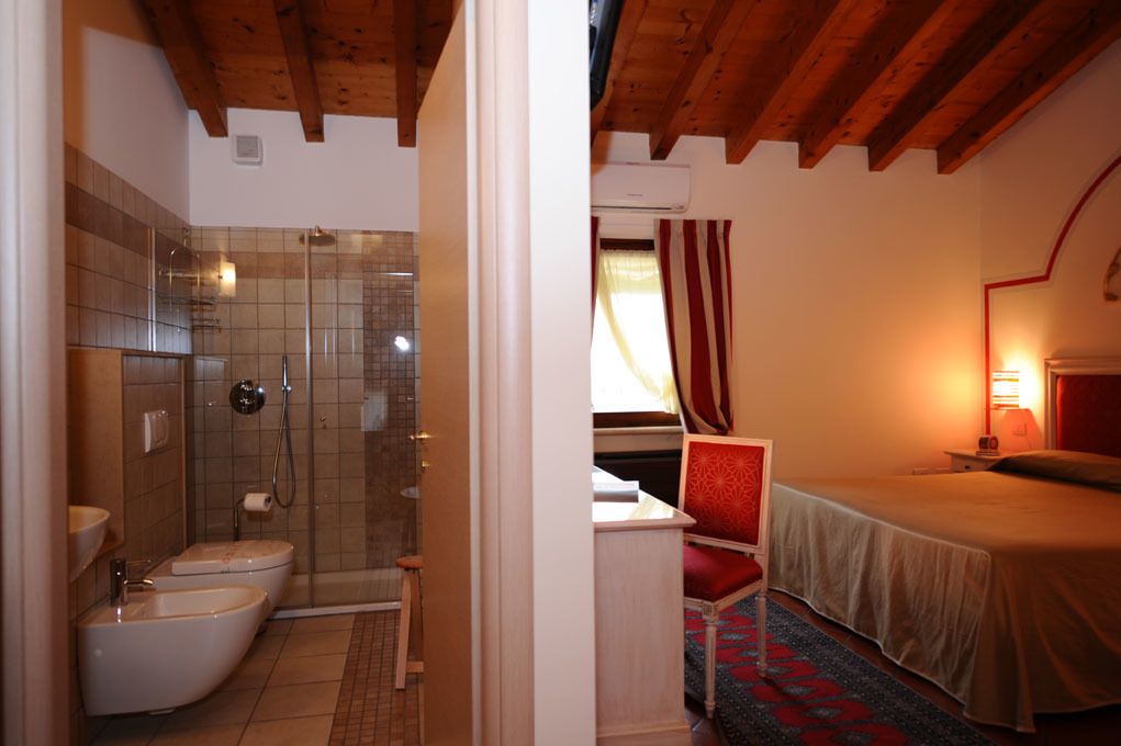 Un baño con ducha, inodoro y lavamanos. Hay una cama, una silla roja y una lámpara.