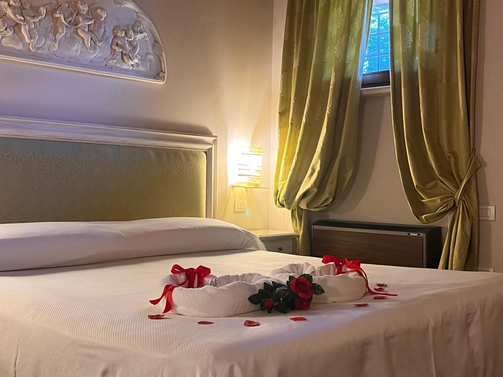 Una cama bien hecha con una almohada decorativa y pétalos de rosa, cabecera con escultura en relieve, lámpara de noche y cortinas verdes.