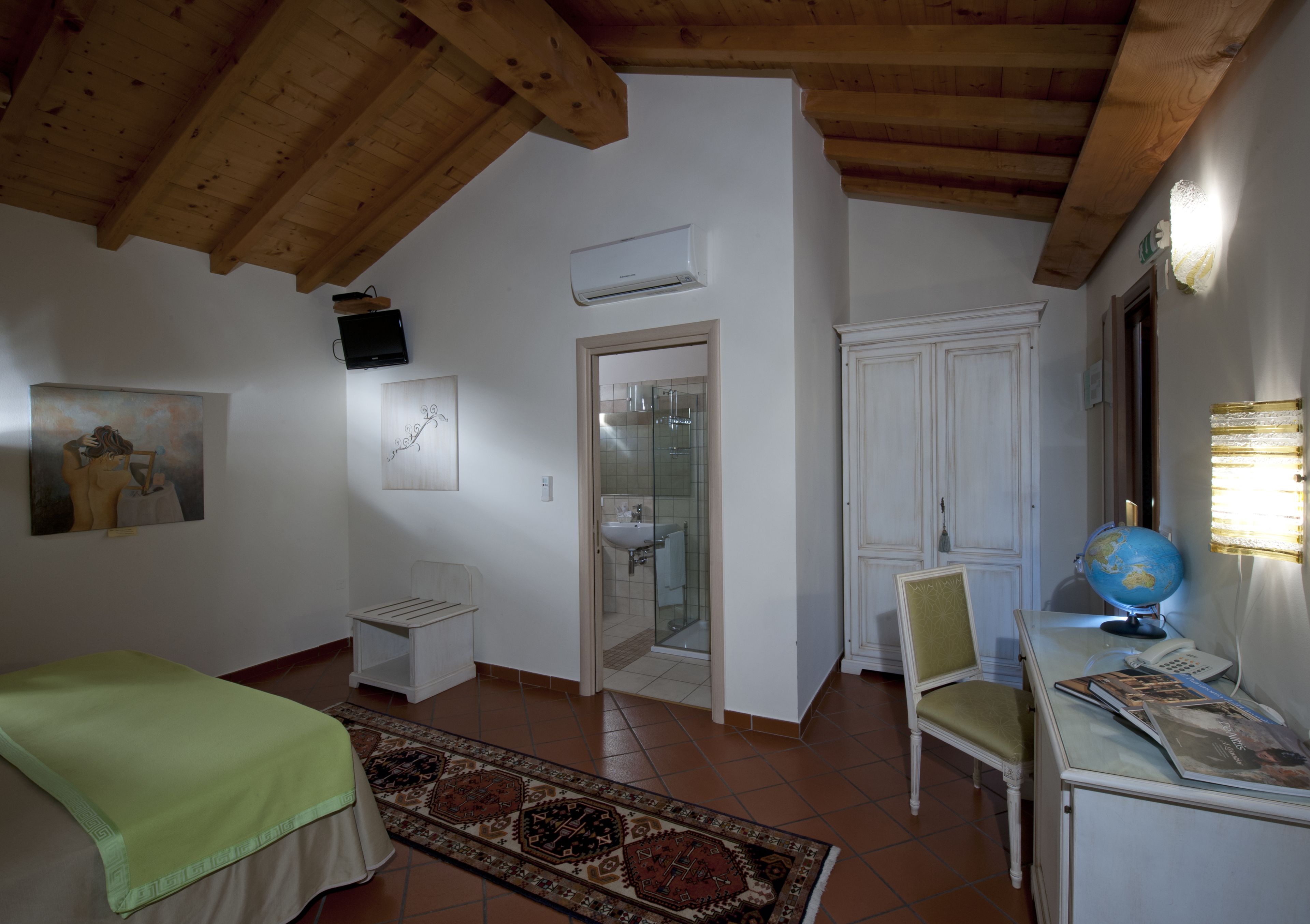 A room with a bed, a desk with a globe, a chair, a painting, and a bathroom door.