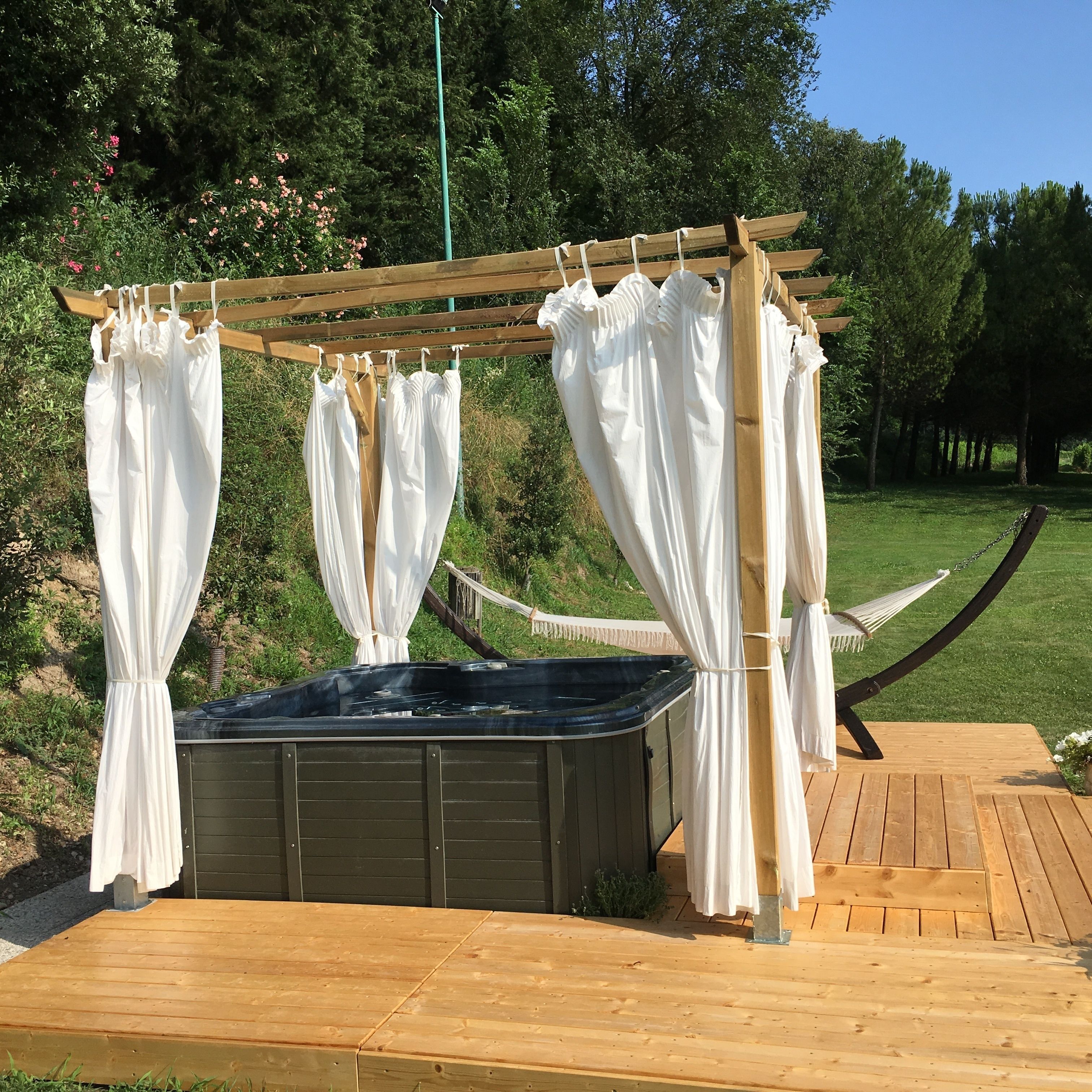 Un bain à remous sous une pergola en bois, agrémentée de rideaux blancs, entouré d’une terrasse en bois et de verdure.