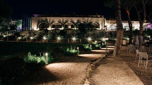 A nighttime view of a building with arched windows and a well-lit pathway leading to it. - Hotel Fontermosa (Orbetello)