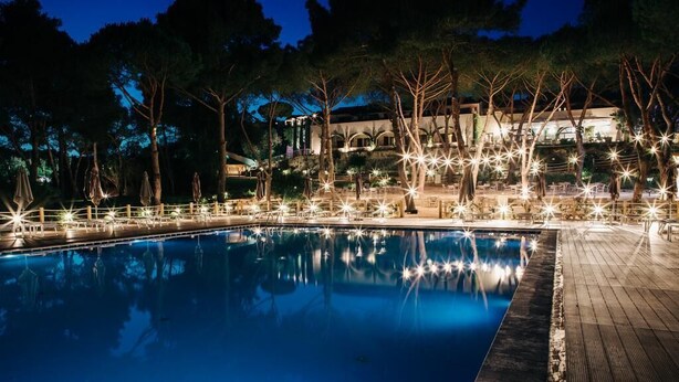 A nighttime pool area with a wooden deck, trees, and a building in the background. - Hotel Fontermosa (Orbetello)